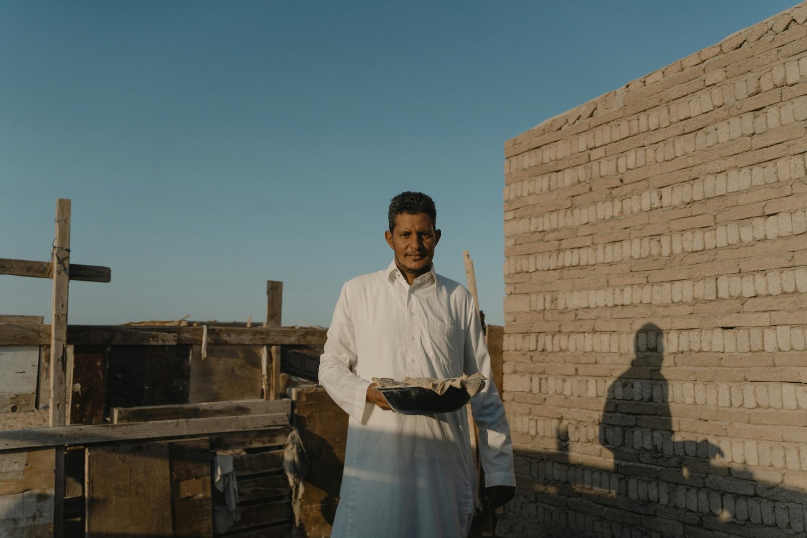 Middle Eastern man in traditional attire holding a bowl, standing outdoors in a rustic setting.